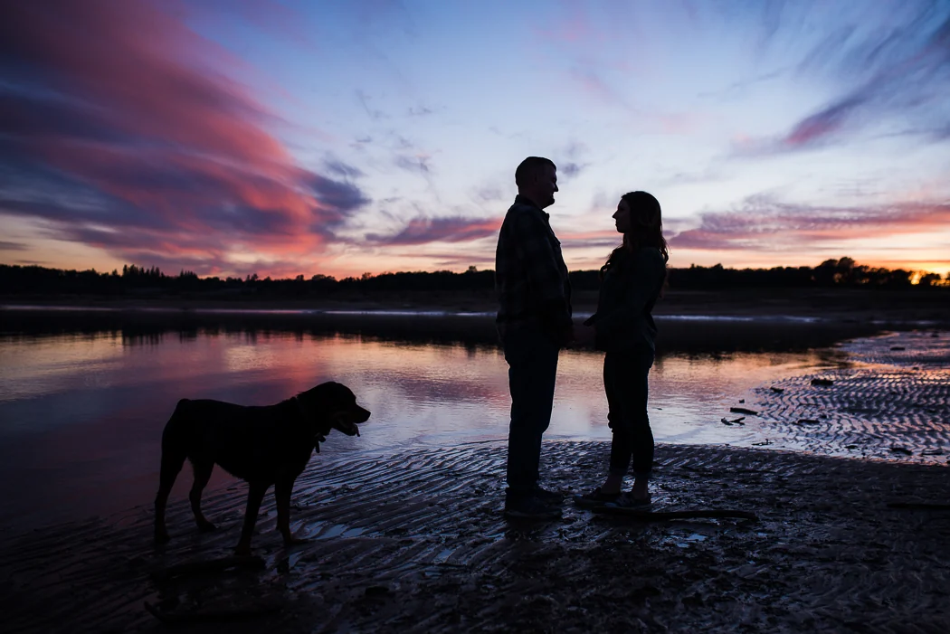 A couple with their dog beside the water at sunset